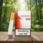 ซองบุหรี่ Journey Original สีขาวแดงวางบนโต๊ะไม้ พร้อมมวนบุหรี่ก้นกรองสีส้ม ฉากหลังเป็นป่าไม้ธรรมชาติ