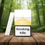 ซองบุหรี่ Marlboro Gold สีขาวทองตั้งตระหง่านคู่กับมวนบุหรี่สีขาวบนโต๊ะไม้ ท่ามกลางบรรยากาศป่าสีเขียว