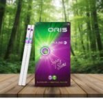 บุหรี่ Oris Pulse Blueberry Menthol ซองสีม่วงเขียวเรืองแสง พร้อมมวนบุหรี่แบบซูเปอร์สลิม 2 เม็ดบีบ วางบนโต๊ะไม้ฉากหลังป่า