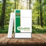 บุหรี่ Atlanta Green ซองแข็งสีขาวคาดแถบสีเขียวและทอง พร้อมมวนบุหรี่สีขาววางบนโต๊ะไม้ในบรรยากาศธรรมชาติ