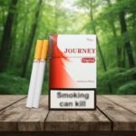 บุหรี่ Journey Original ซองสีขาวคาดแถบสีแดงดีไซน์โฉบเฉี่ยว พร้อมมวนบุหรี่ก้นกรองสีส้มวางบนโต๊ะไม้ฉากหลังป่า