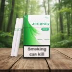 บุหรี่ Journey Green ซองแข็งสีขาวคาดแถบกราฟิกสีเขียวสดใส พร้อมมวนบุหรี่สีขาววางบนโต๊ะไม้ในบรรยากาศป่าไม้