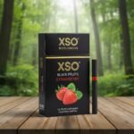ซองบุหรี่ XSO Strawberry สีดำดีไซน์หรูหราพร้อมรูปสตรอว์เบอร์รี วางคู่กับมวนบุหรี่สีดำบนโต๊ะไม้ฉากหลังป่าธรรมชาติ