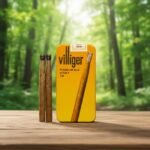 กล่องอลูมิเนียมซิการ์ Villiger Honey Tip สีเหลืองส้มวางบนพื้นไม้ มีมวนซิการ์ใบยาสูบธรรมชาติวางคู่กันในบรรยากาศป่าไม้