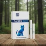 บุหรี่ Raison Blue ซองสีขาวลายแมวสีน้ำเงินสไตล์กราฟิก วางคู่กับมวนบุหรี่บนโต๊ะไม้ฉากหลังป่า