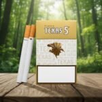 บุหรี่ Texas 5 Gold ซองสีทองลายใบยาสูบ วางคู่กับมวนบุหรี่บนโต๊ะไม้ฉากหลังป่าสีเขียว