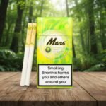 บุหรี่ Mars Honeydew ซองสีเขียวสว่างลายกราฟิก วางคู่กับมวนบุหรี่บนโต๊ะไม้ฉากหลังป่า