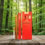 ซิการ์ Villiger Red Tube Vanilla ในหลอดอลูมิเนียมสีแดงและซองสีแดงข้างมวนซิการ์ใบยาสูบธรรมชาติ วางบนโต๊ะไม้ฉากหลังป่าสีเขียว