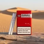 ซองบุหรี่ Dunhill Red 10mg สีแดงสดใส พร้อมมวนบุหรี่ วางบนขอนไม้ท่ามกลางวิวทะเลทรายสีทอง